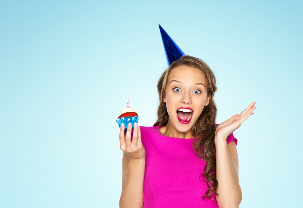 emotion, expression and celebration concept - happy young woman or teen girl in pink dress and party cap with birthday cupcake over blue background. happy woman or teen girl with birthday cupcake