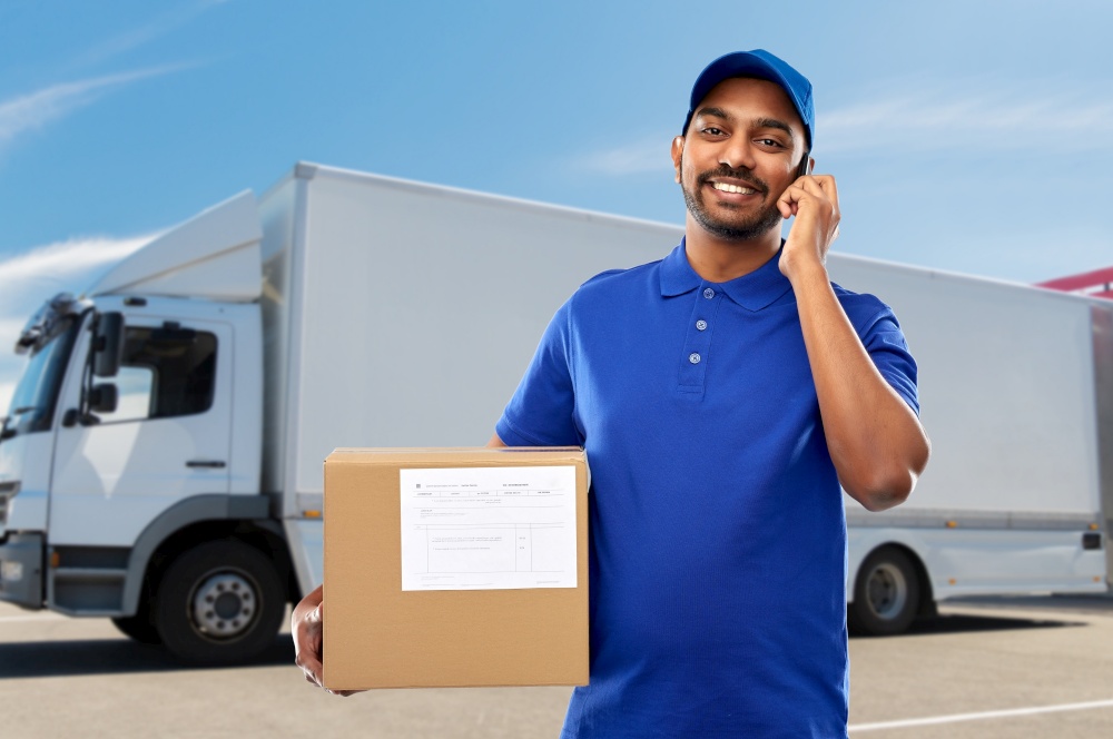 mail service, communication and shipment concept - happy indian delivery man with smartphone and parcel box in blue uniform over truck on street background. indian delivery man with smartphone and parcel box