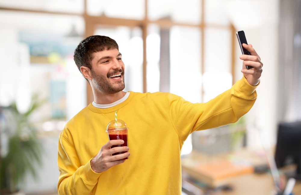 technology and people concept - happy smiling young man taking selfie with smartphone with tomato juice in takeaway plastic cup with paper straw over office background. happy man with smartphone and juice taking selfie
