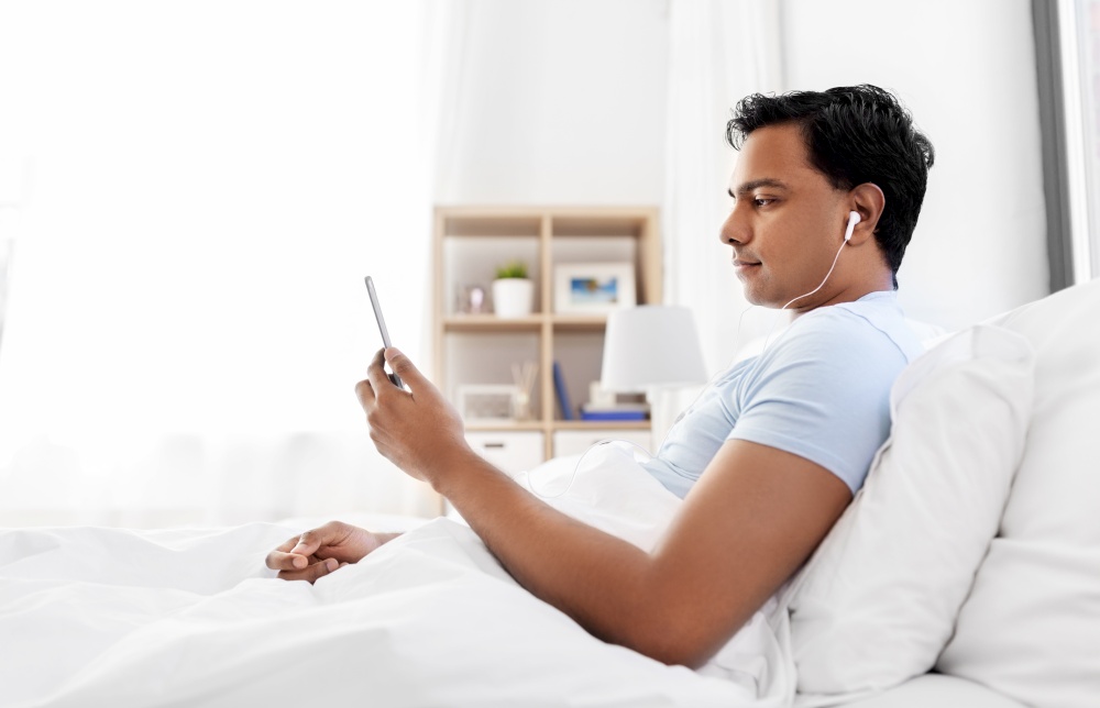 technology, bedtime and rest concept - indian man in earphones with smartphone lying in bed and listening to music at home. indian man in earphones with phone in bed at home