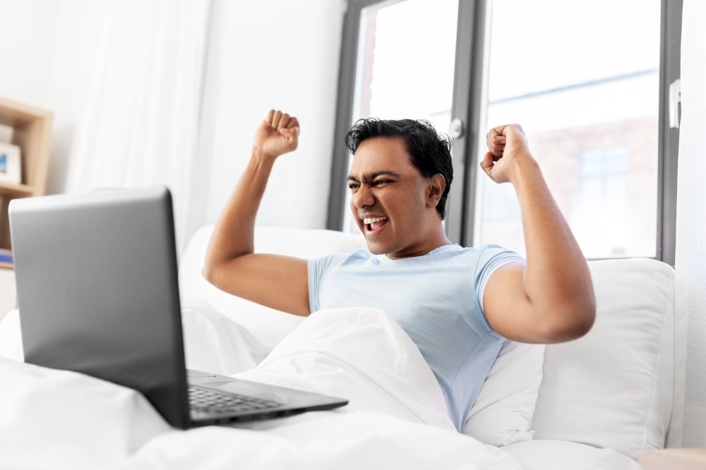 people, technology and success concept - happy smiling indian man with laptop computer lying in bed and making winning gesture at home. happy indian man with laptop in bed at home