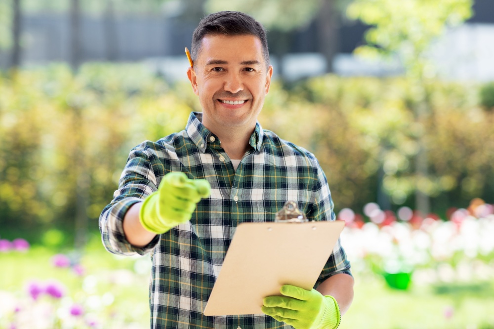 gardening and people concept - happy smiling middle-aged man with clipboard pointing finger to camera at summer garden. happy smiling man with clipboard at summer garden
