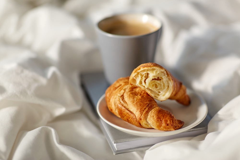 morning, hygge and breakfast concept - croissants, cup of coffee and book in bed at home. croissants, cup of coffee and book in bed at home