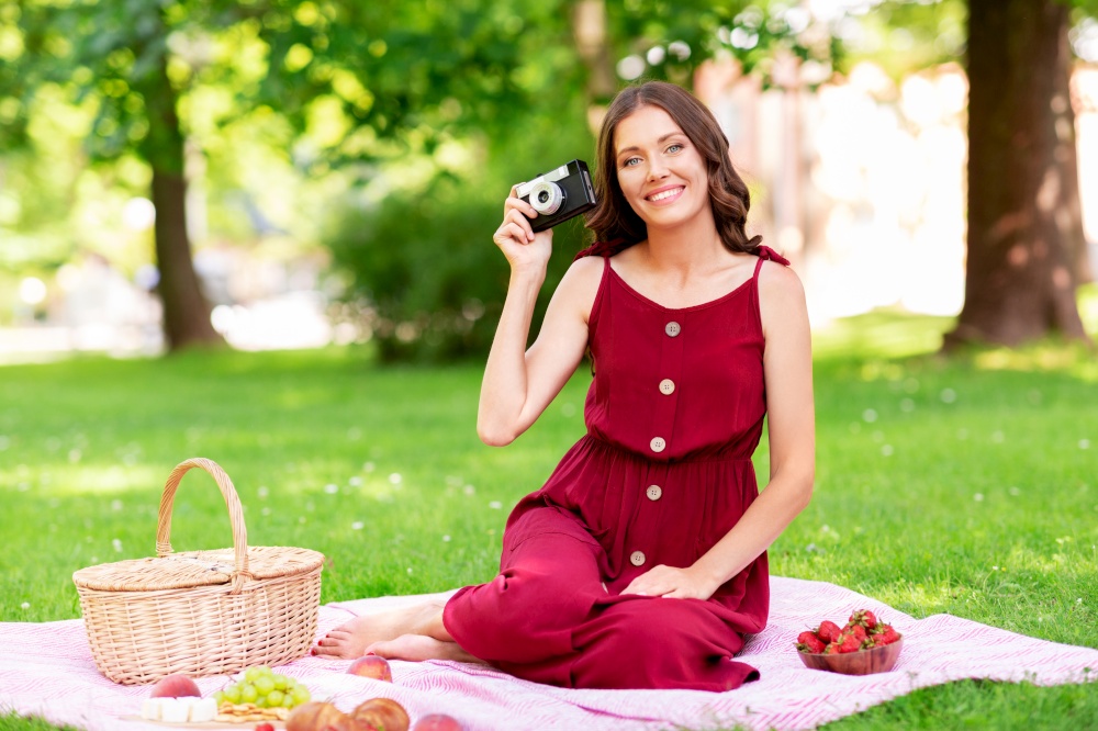 photography, leisure and people concept - happy smiling woman with camera, picnic basket sitting on blanket at summer park. happy woman with camera on picnic at park
