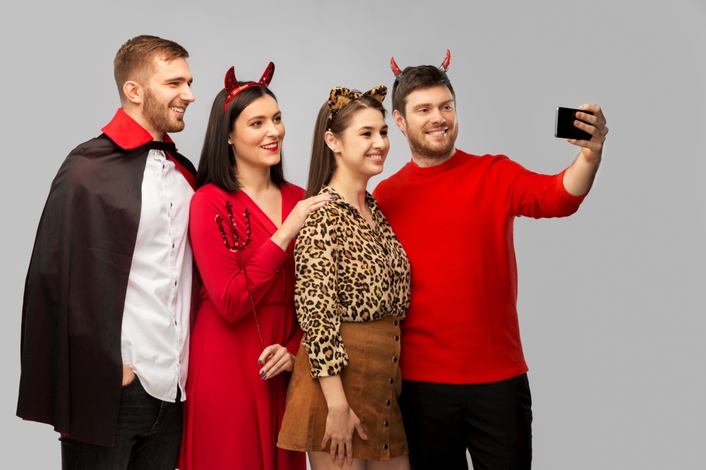 friendship, holiday and people concept - group of happy smiling friends in halloween costumes of vampire, devil and cheetah taking selfie by smartphone over grey background. happy friends in halloween costumes taking selfie