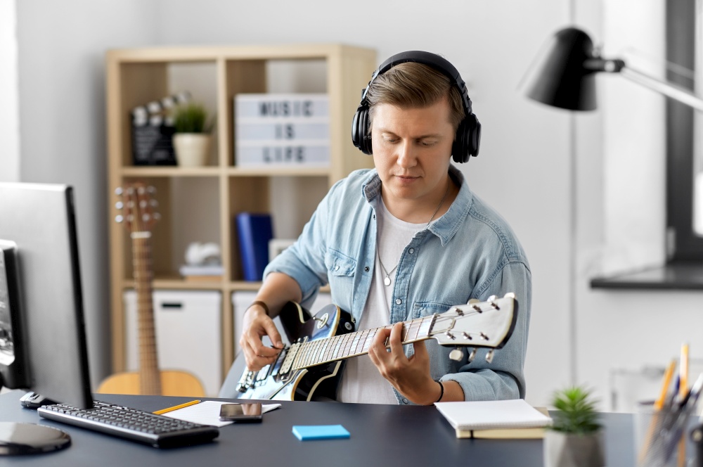 leisure, music and people concept - young man or musician in headphones with computer playing bass guitar sitting at table at home. man in headphones playing bass guitar at home