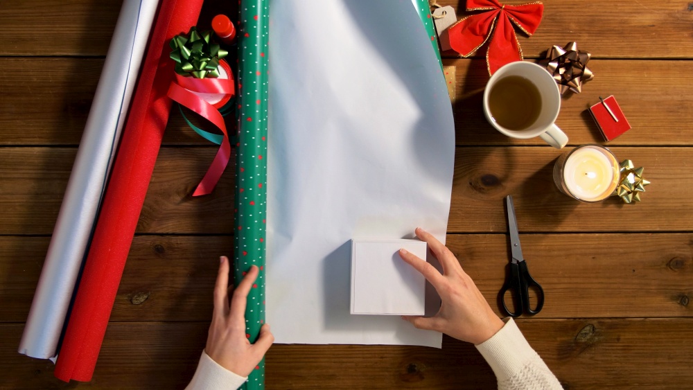 holidays, new year and christmas concept - hands wrapping gift box into green paper on wooden table. hands wrapping christmas gift into paper at home