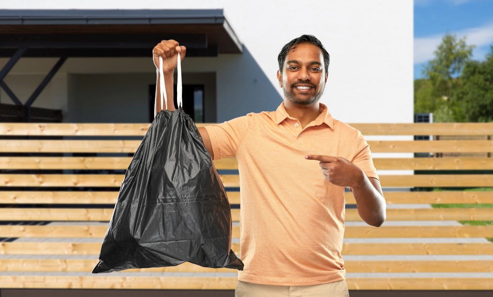 recycling, sorting and sustainability concept - smiling young indian man holding trash bag over house background. smiling indian man holding trash bag