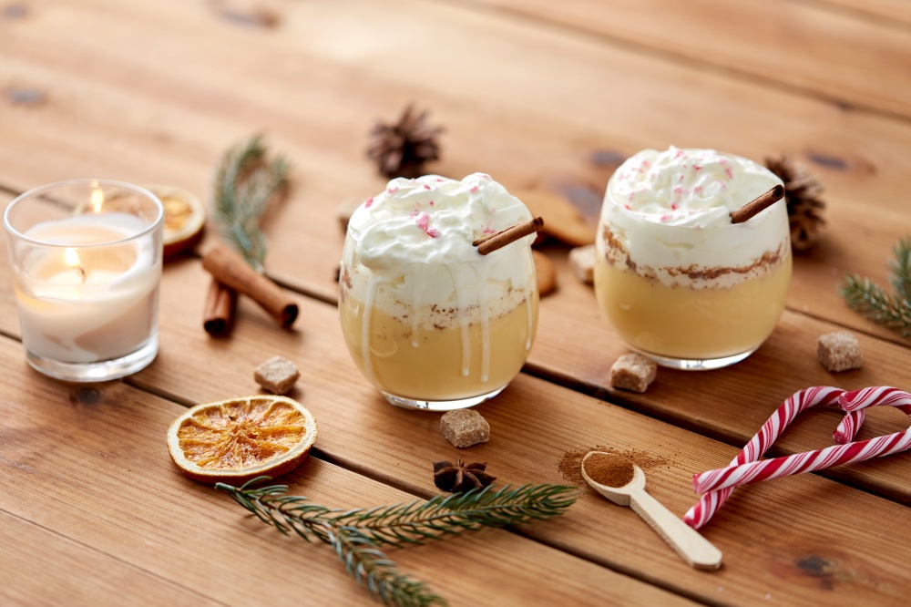 christmas and seasonal drinks concept - glasses of eggnog with whipped cream topping and cinnamon, fir tree branches, brown sugar and candle burning on wooden background. glasses of eggnog with whipped cream and spices