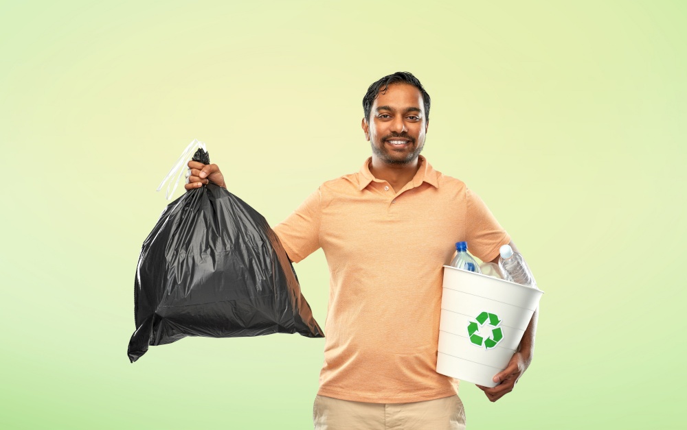recycling, sorting and sustainability concept - smiling young indian man holding bucket with plastic bottles and trash bag over green background. smiling indian man sorting paper and plastic waste