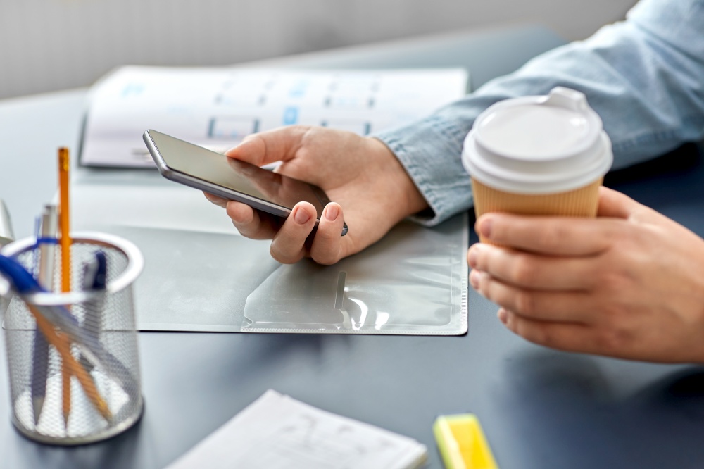 business, technology and people concept - close up of hands using smartphone and drinking takeaway coffee from paper cup at office. woman with coffee using smartphone at office