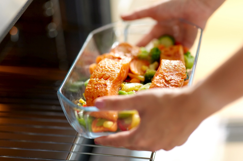 culinary, food and people concept - woman cooking salmon fish with vegetables in baking dish in oven at home kitchen. woman cooking food in oven at home kitchen