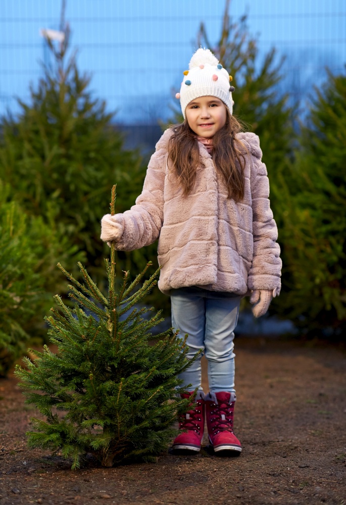 winter holidays and people concept - happy smiling little girl choosing christmas tree at street market in evening. little girl choosing christmas tree at market