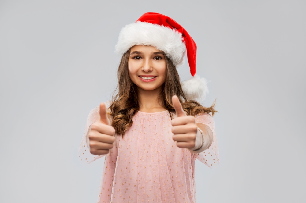christmas, holidays and people concept - happy smiling teenage girl in santa helper hat showing thumbs up over grey background. happy teenage girl in santa hat showing thumbs up