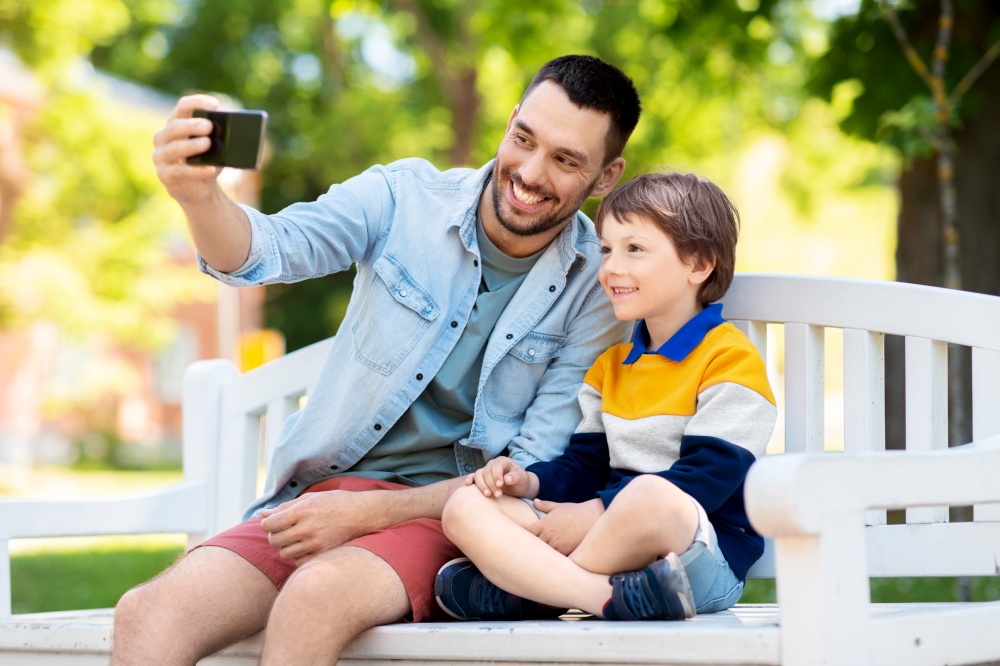 family, fatherhood and people concept - happy father with little son taking selfie picture with smartphone sitting on bench at summer park. father and son taking selfie with phone at park