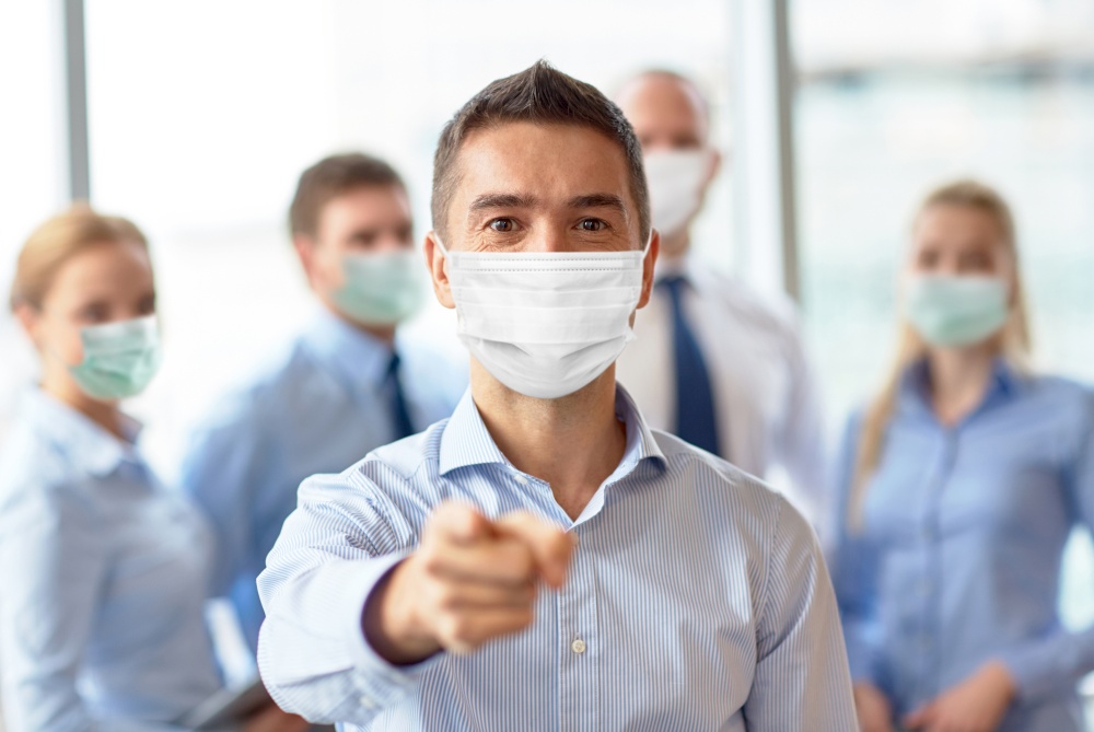 business, people and pandemic concept - businessman wearing face protective medical mask for protection from virus disease pointing finger to camera at office. businessman in mask pointing finger at office