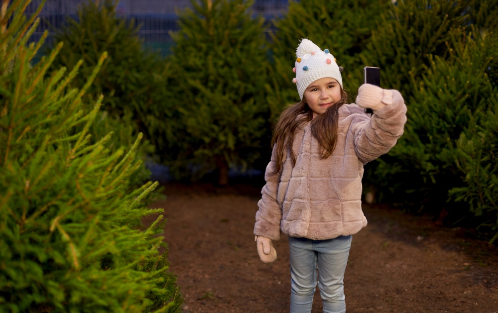 winter holidays, children and people concept - happy smiling little girl with smartphone taking selfie at christmas tree market in evening. little girl taking selfie at christmas tree market