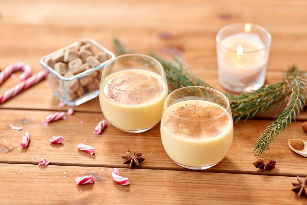christmas and seasonal drinks concept - glasses of eggnog with candy canes, sugar, fir tree branches and candle burning on wooden background. glasses of eggnog, ingredients and spices on wood