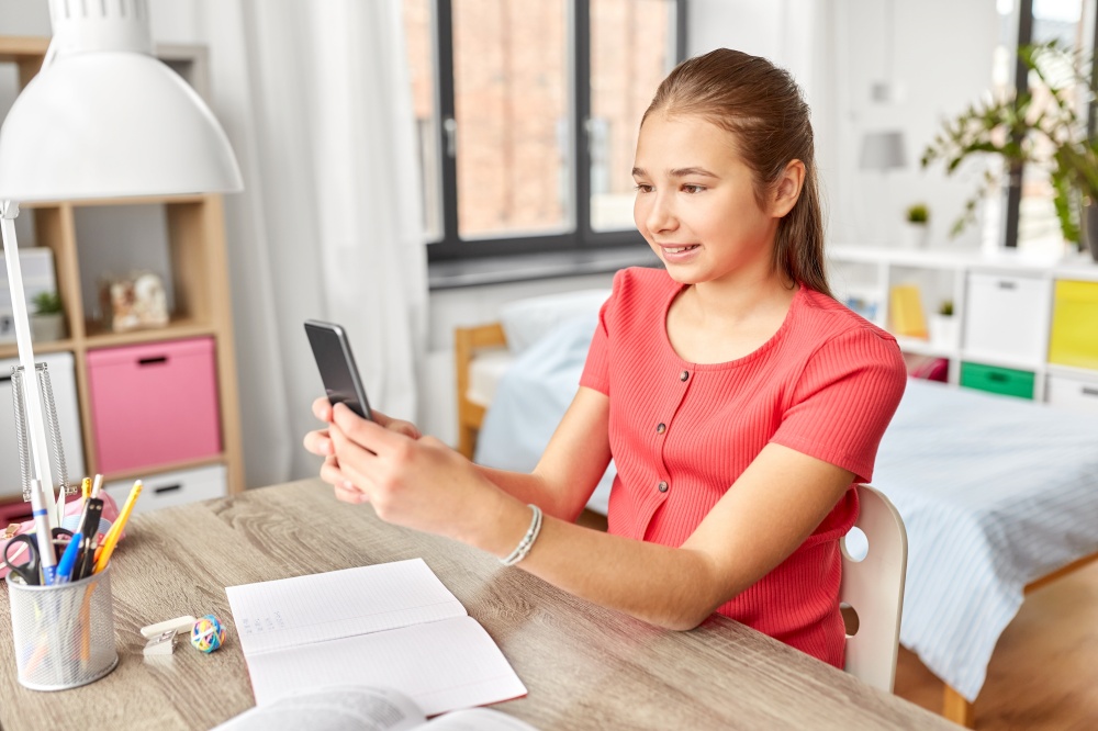children, technology and communication concept - happy smiling teenage student girl distracting from homework and texting on smartphone at home. girl with smartphone distracting from homework