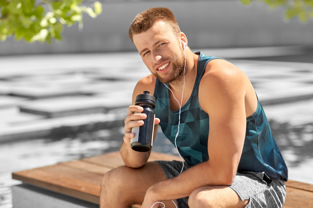 fitness, sport and people concept - happy smiling young man with earphones and bottle of water sitting on city bench. sportsman with earphones and bottle in city