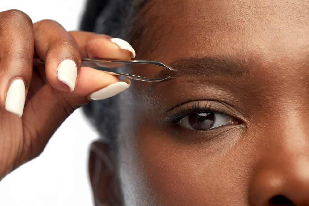grooming and people concept - close up of face of young african american woman with tweezers tweezing her eyebrow over white background. african woman with tweezers tweezing her eyebrow