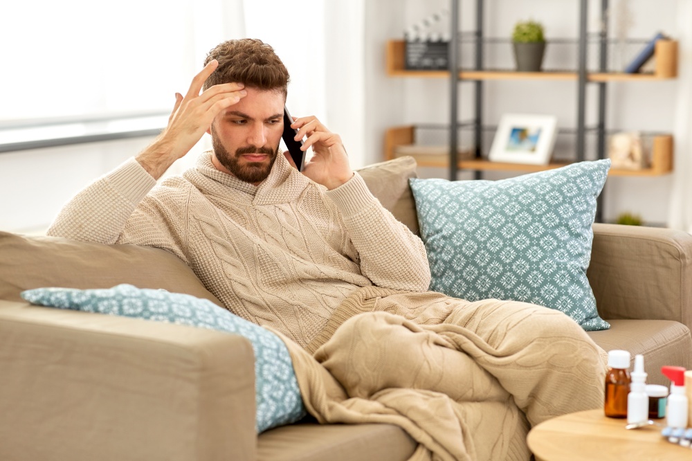 health, cold and people concept - sick young man in blanket with smartphone and medicine calling doctor at home. sick young man calling on smartphone at home