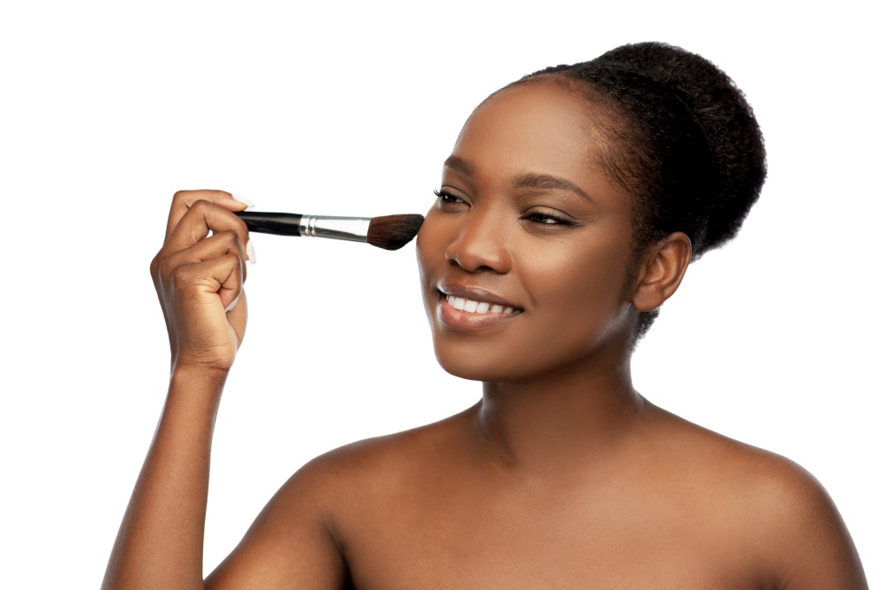 beauty, cosmetics and people concept - portrait of happy smiling young african american woman with make up brush applying blush to her face over white background. woman with make up brush applying blush to face