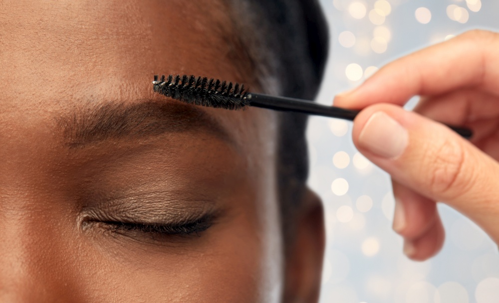 beauty, make up and cosmetics concept - close up of face of young african american woman and hand with mascara brush applying eyebrow shadows over festive lights background. face of african woman and hand with mascara brush