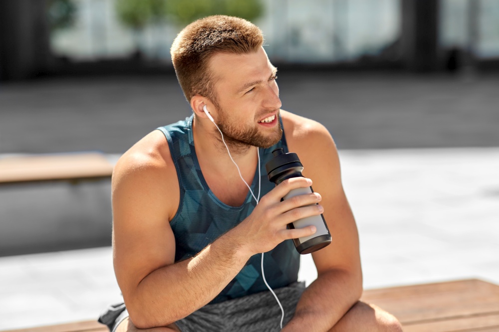 fitness, sport and people concept - young man with earphones and bottle of water sitting on city bench. sportsman with earphones and bottle in city