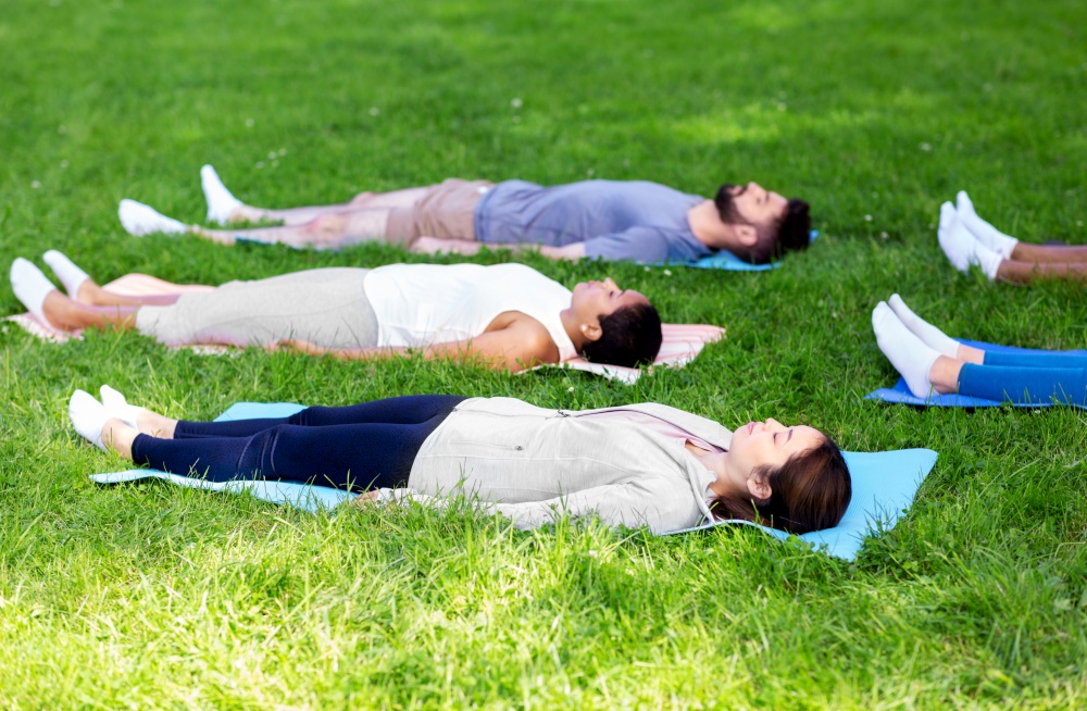 fitness, sport and healthy lifestyle concept - group of happy people doing yoga corpse pose or savasana at summer park. group of people doing yoga at summer park