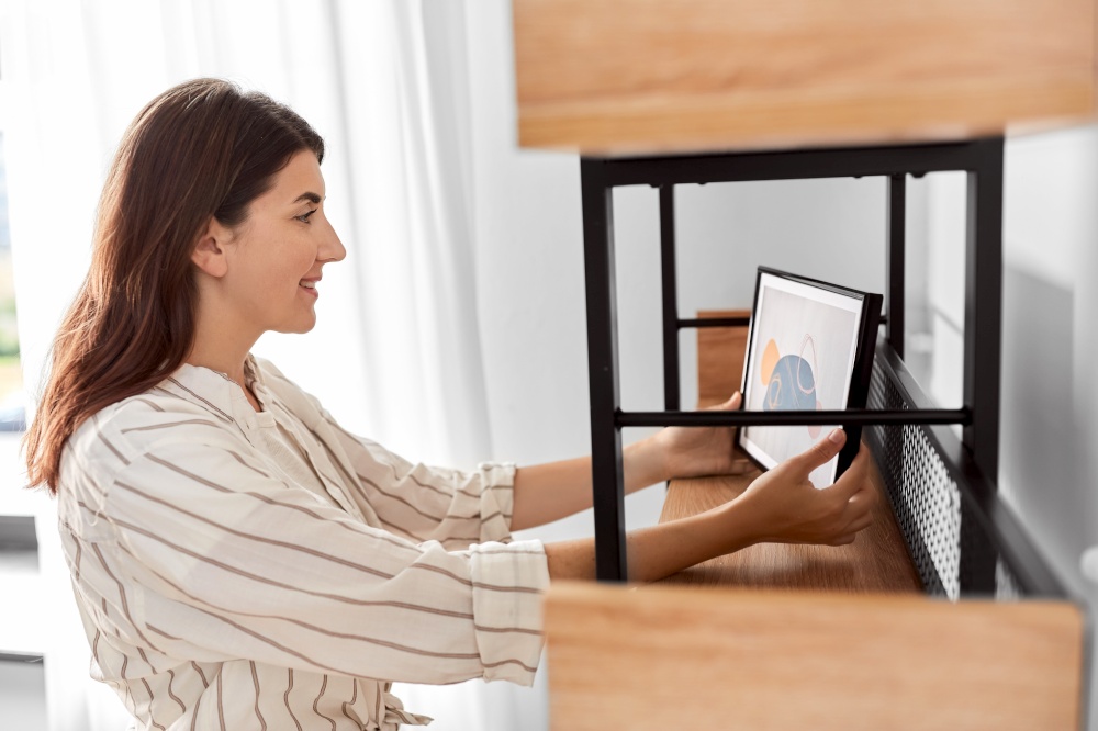 home improvement, decoration and people concept - happy smiling woman placing picture in frame to shelf. woman decorating home with picture in frame