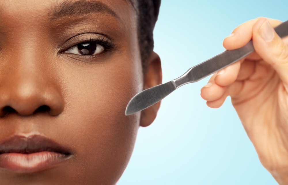 beauty, plastic surgery and people concept - close up of face of beautiful young african american woman and hand with scalpel knife over blue background. face of african american woman and scalpel knife