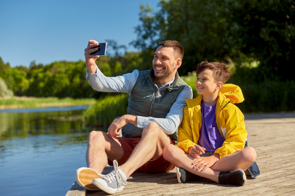 family, generation and technology concept - happy father and son taking selfie with smartphone on river berth. father and son taking selfie with phone on river