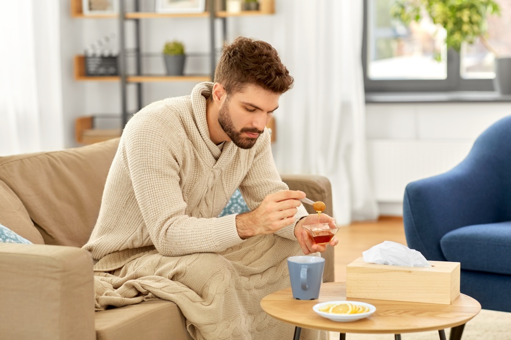 health, cold and people concept - sick young man in blanket drinking hot tea with lemon and honey at home. sick young man in blanket drinking hot tea at home