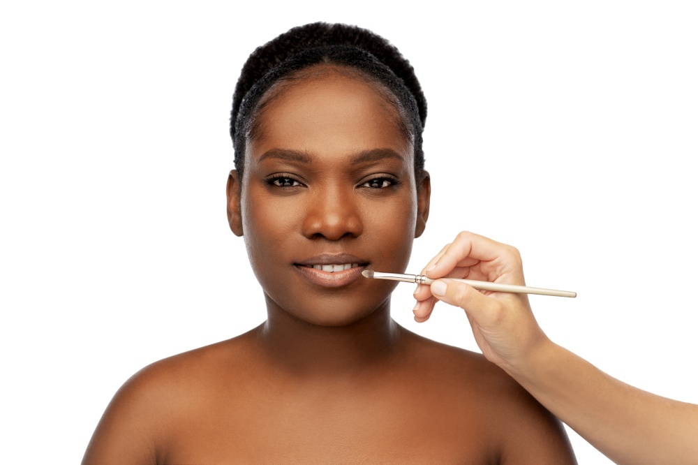 beauty, cosmetics and people concept - close up of face of beautiful smiling young african american woman and hand with make up brush applying lipstick or gloss to lips over white background. face of african woman and hand with make up brush