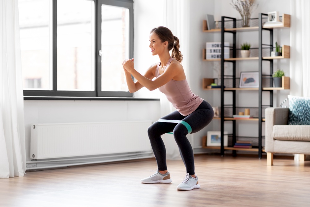 sport, fitness and healthy lifestyle concept - smiling young woman with resistance band exercising and doing squats at home. woman exercising with resistance band at home