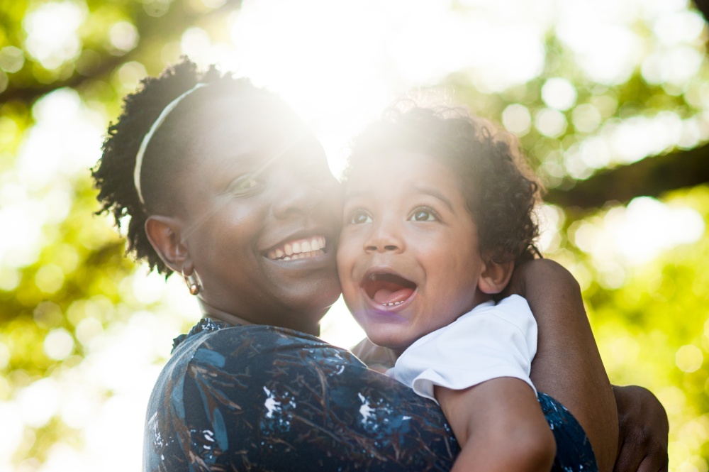 African mother smiling to the camera  holding her toddler boy in her arms.  The sunlight is illuminating the scene. Natural Flare