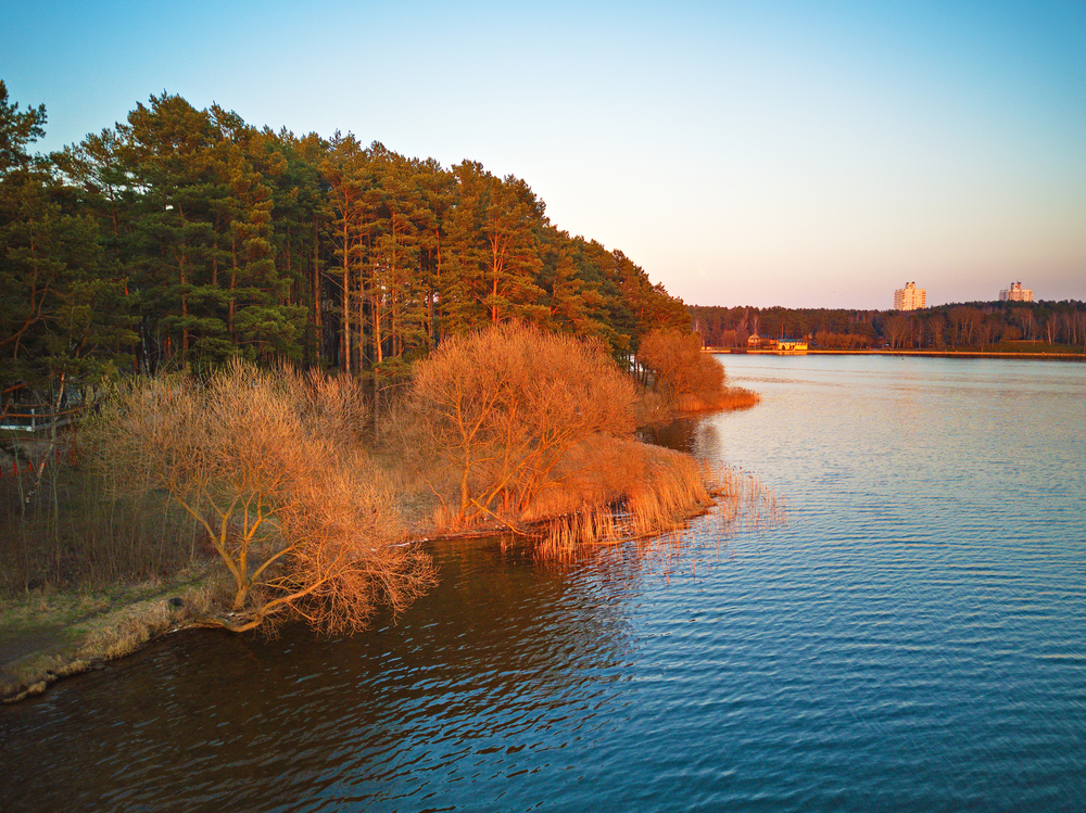 Panoramic landscape of lake shore with colorful trees in spring sunlight. April evening at the lake near Minsk, Belarus. Recreation zone aerial view. Beautiful sunset scene.