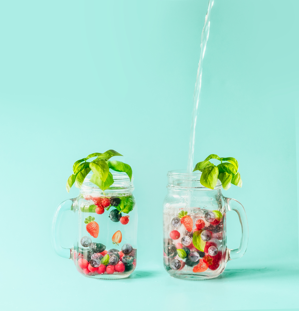 Stream of fresh water pours into a bottle with berries detox fruit infused water in Mason jar flavored with herb leaves at sunny turquoise blue background. Summer mood.  Healthy drinks and lifestyle.