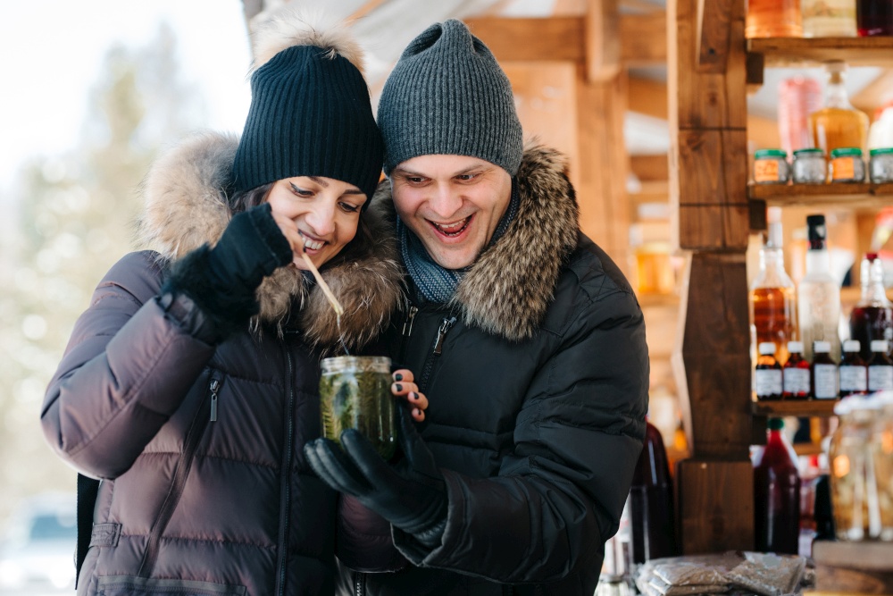 joyful girl and guy in warm winter clothes at the market try honey and drinks