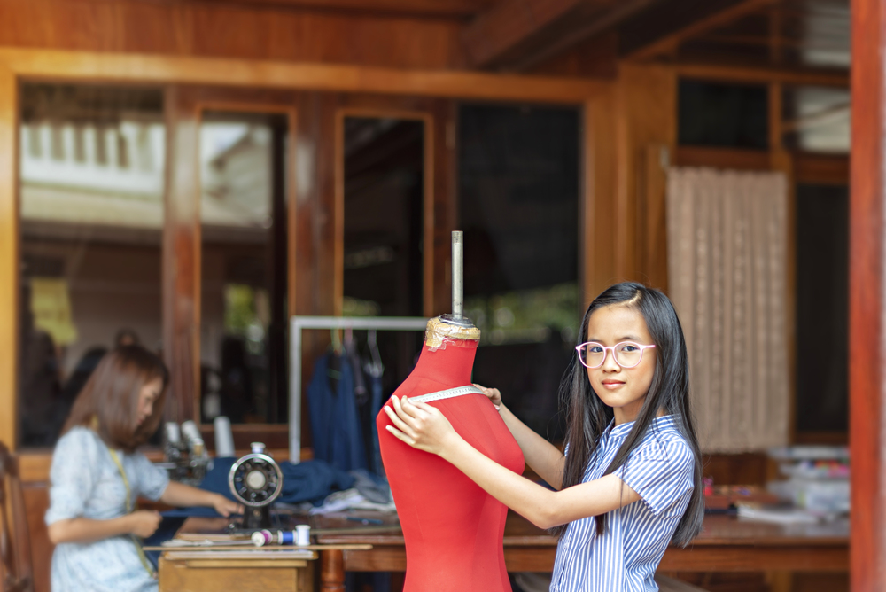 asian little girl dressmaker measuring the  dummy and looking at camera with smile while standing in workshop. asian little girl dressmaker