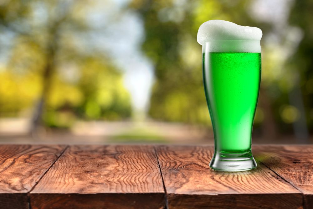 Glass of fresh cold green beer with extra foam on a wooden table with blurred green summer leaves on natural background with bokeh, copy space. Happy St.Patrick &rsquo;s Day concept.. Green beer in glass on wooden table against blurred park.