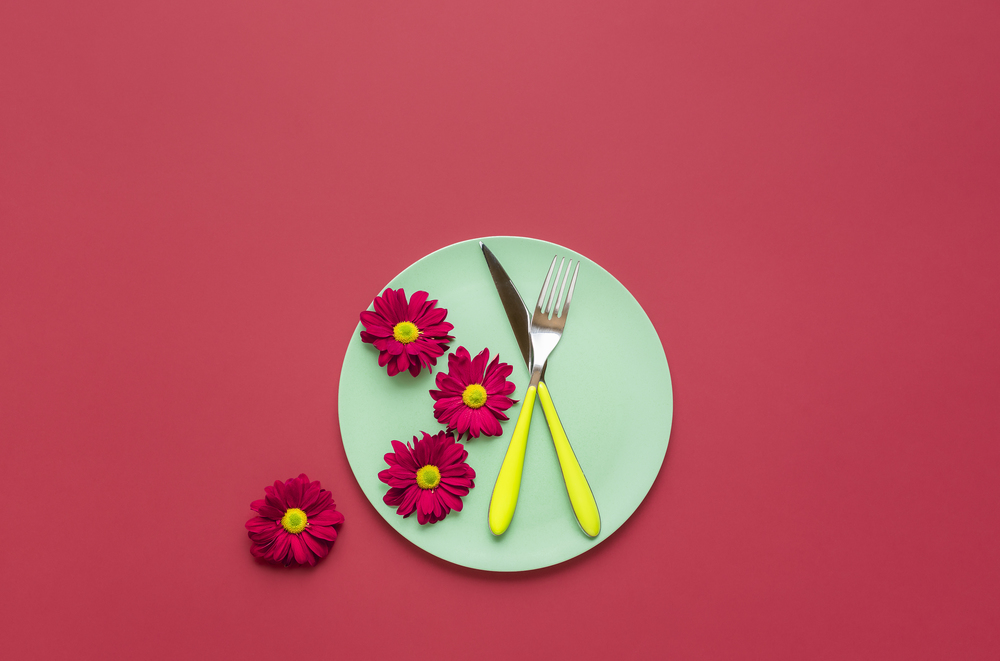 Green dish with cutlery and red flowers, on a red background. Above view of spring table settings. Healthy food context. Dinner table arrangement.