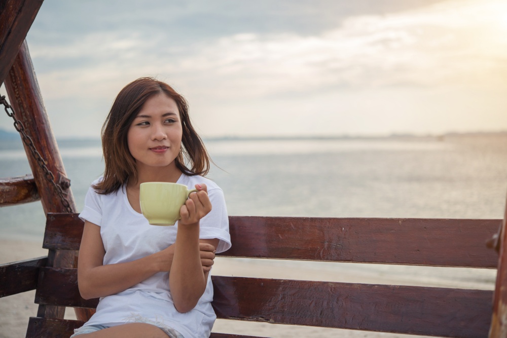 Beautiful woman drink coffee while sitting on swing on the beach.
