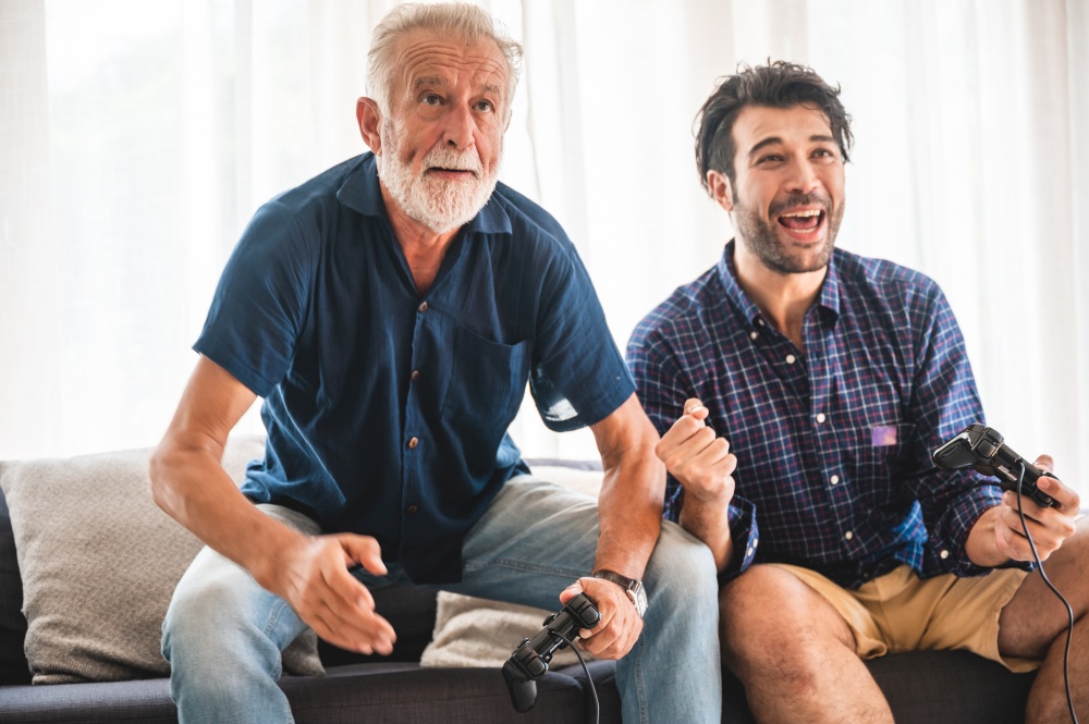 Happy two age generations men family old father embracing young grown adult son having fun enjoying video game