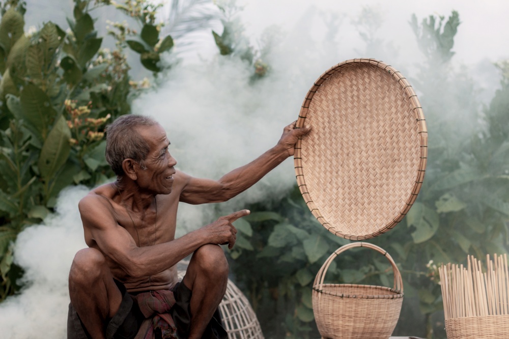 Asian old man is showing a bamboo tray in the countryside.