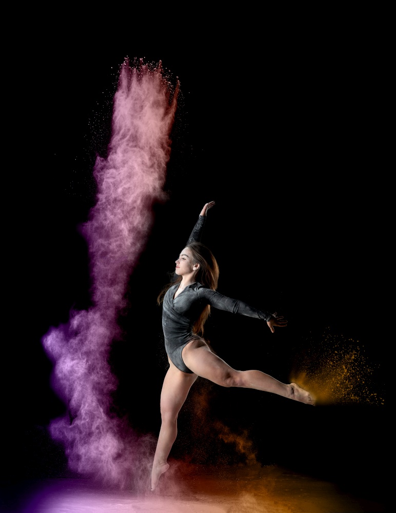 beautiful young caucasian woman in a black bodysuit with a sports figure dancing in a purple-yellow cloud of flour on a black background, explosion and expression in motion