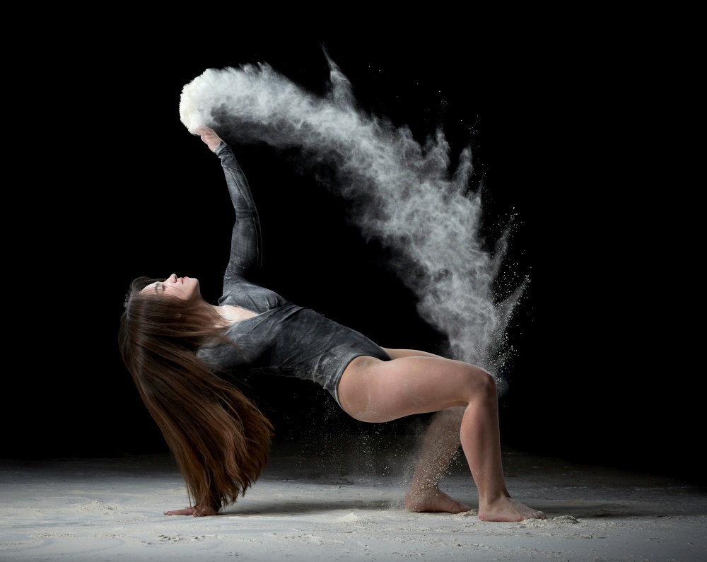 young beautiful woman with long hair is dressed in a sports black bodysuit and sits on the floor and throws white flour over her, black background