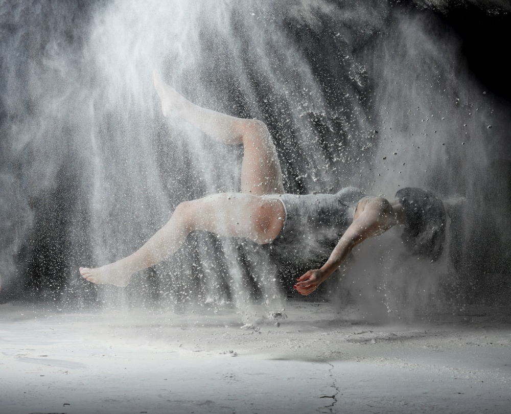 beautiful young woman in a black bodysuit with a sports figure soars on a black background in a spray of white flour, creative photo