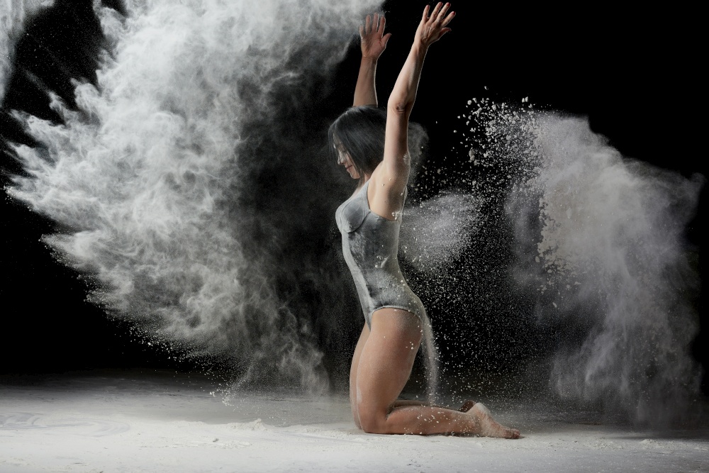 young woman with an athletic body sits on the floor and throws white flour up, dust flies in different directions, an athlete is dressed in a black bodysuit